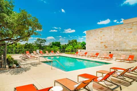 Modern pool area with bright orange lounge chairs, stone building, green trees, and clear blue sky.