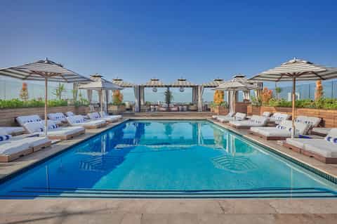 Rooftop pool with striped umbrellas, lounge chairs, curtained cabanas, and city skyline views