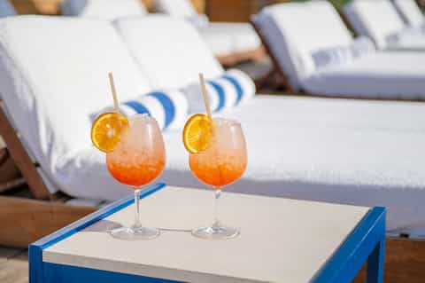 Two coral-colored cocktails with orange slice garnishes on poolside table and lounge chairs