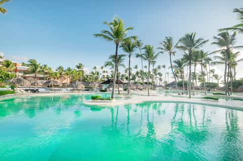Tropical resort pool with crystal blue water, palm trees, and thatched-roof beach structures