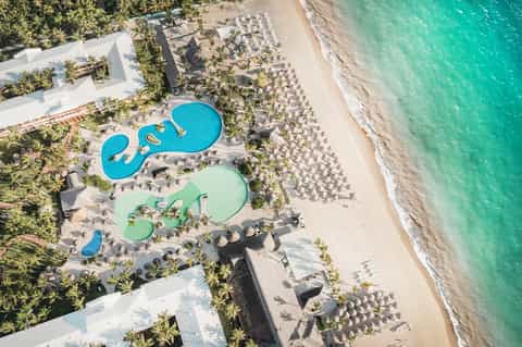 Aerial view of beachfront resort with blue pools, palm trees, white umbrellas, and turquoise ocean