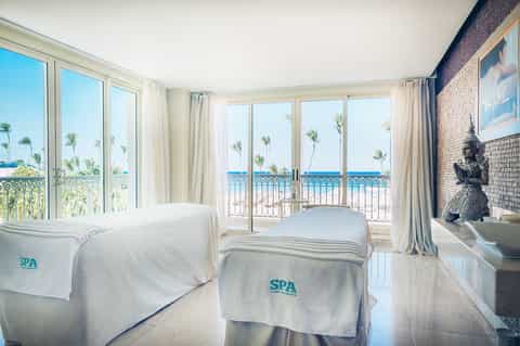 Spa massage treatment room with ocean and palm tree views through glass doors, two massage beds with white linens