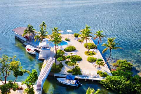 Aerial view of private island resort with palm trees, white sand beach, boats, and turquoise water