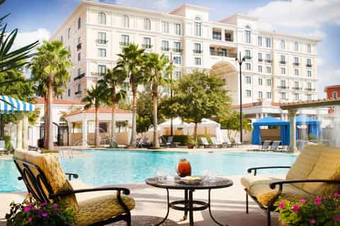 Resort pool with cream building, palm trees, white umbrellas, and lounge seating