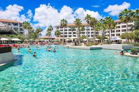 Crowded lagoon-style pool at resort with families swimming, palm trees, and multi-story hotel building
