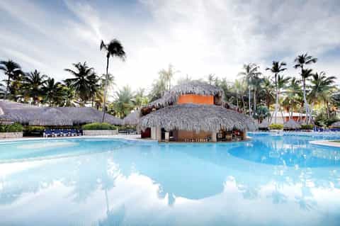 Large resort pool with thatched-roof bar structure and palm trees at sunset