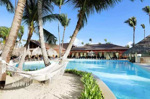 Tropical resort pool surrounded by palm trees, hammocks, and thatched-roof buildings