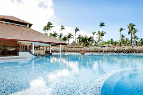 Lagoon-style resort pool with thatched cabanas, palm trees, and modern building with wooden architecture
