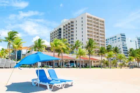 Sandy beach with blue loungers, blue umbrella, high-rise hotel buildings, and palm trees