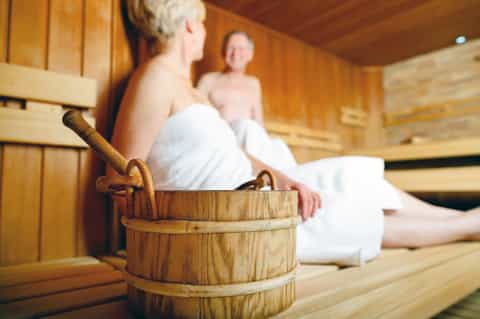 Wooden sauna interior with two people wrapped in white towels on wooden benches.