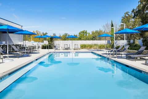 Resort swimming pool with blue umbrellas, lounge chairs, and white privacy fence