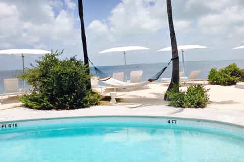 Beachfront pool with white umbrellas, hammocks between palm trees, and ocean view