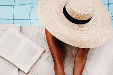 Person poolside wearing oversized straw hat, reading book by turquoise water