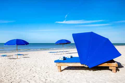 Sandy beach with blue umbrellas, lounge chairs, and clear sky with calm ocean in background