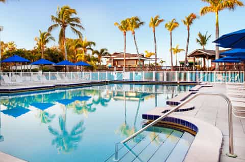 Resort pool with palm trees, blue umbrellas, white fence, and tropical clubhouse architecture