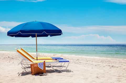 Beach chairs under blue umbrella with vintage suitcase on sandy beach overlooking calm ocean