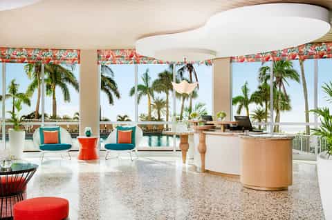 Bright, open lounge with tropical views, palm trees through windows, teal seating, orange accent table, and modern bar