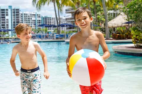 Two young boys playing in resort pool with beach ball and tropical palm tree landscaping