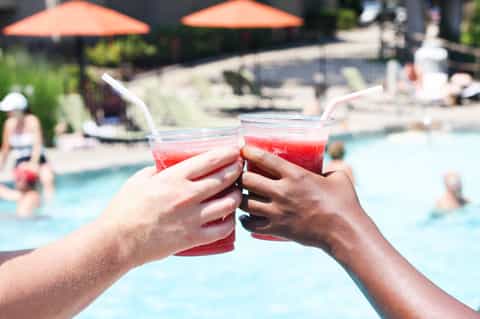 Two people clinking red cocktail drinks by a pool with orange umbrellas and swimmers in the background