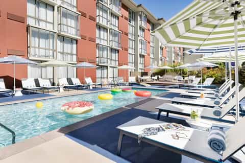 Modern resort pool with colorful floating pastry inflatables, striped umbrellas, and coral building
