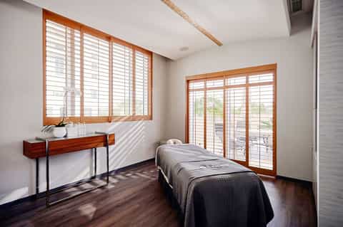 Bright spa treatment room with massage bed and ocean view through shuttered windows