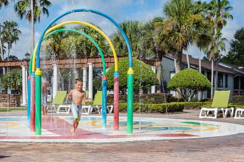Colorful splash pad with blue and yellow arches, child playing, palm trees, resort building
