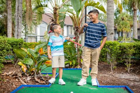 Two young boys playing mini golf outdoors surrounded by tropical plants and palm trees