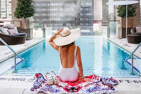 Woman in white swimsuit and hat sitting at rooftop pool overlooking city skyline
