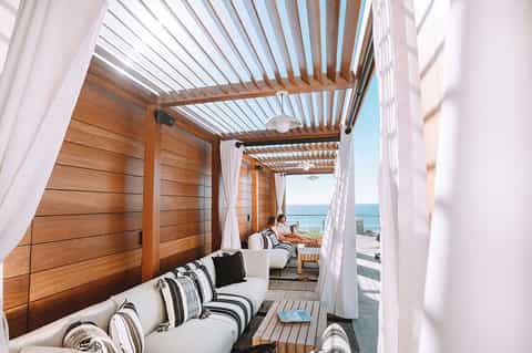 Modern wooden pergola lounge area overlooking the ocean with comfortable seating and white curtains