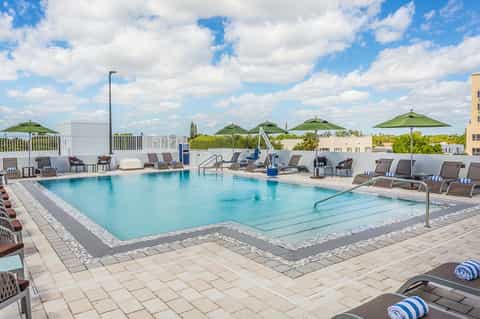 Outdoor swimming pool with lounge chairs, green umbrellas and landscaped resort grounds under blue sky