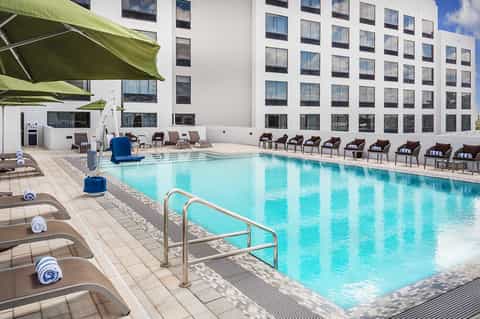 Large outdoor pool with turquoise water, lounge chairs with rolled towels, and modern hotel building