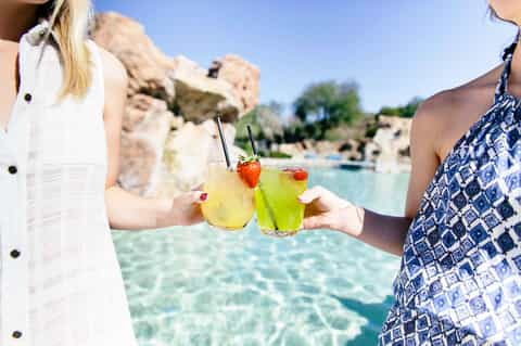 Two women toasting with colorful cocktails by a turquoise lagoon with rocky cliffs