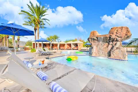 Resort pool with rock water feature, palm trees, blue umbrellas, and cabanas under bright sunny sky