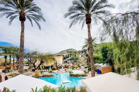 Curved resort pool with palm trees, white umbrellas, and mountain backdrop at dusk