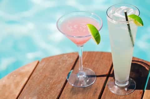 Two tropical cocktails with lime garnish on wooden deck overlooking turquoise water