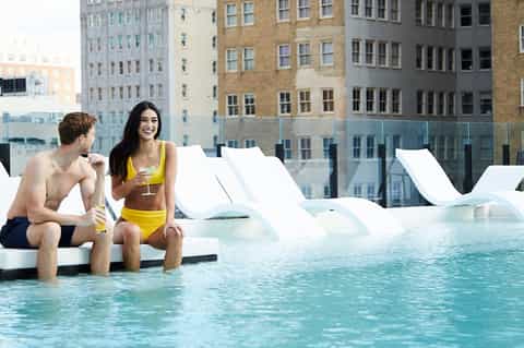 Couple relaxing at the edge of a rooftop infinity pool with white loungers overlooking urban cityscape