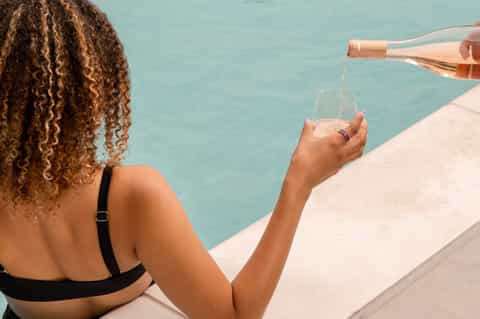 Woman pouring champagne by turquoise pool edge on sunny resort day