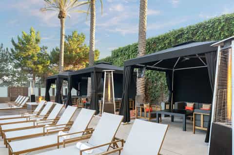 Resort poolside cabanas with black curtains, lounge chairs, and palm trees at sunset