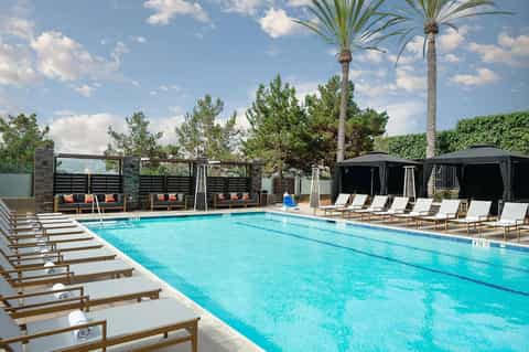 Resort swimming pool with blue lounge chairs, black cabanas, palm trees, and mountain backdrop