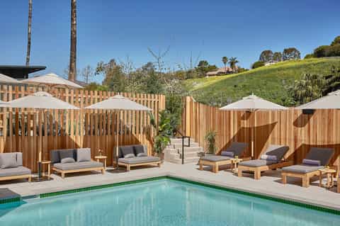 Resort pool deck with turquoise water, wooden lounge chairs, white umbrellas, and wooden privacy fence backdrop