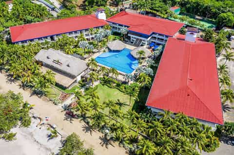 Aerial view of beachfront resort with red roof, blue pool, palm trees, and sandy beach access