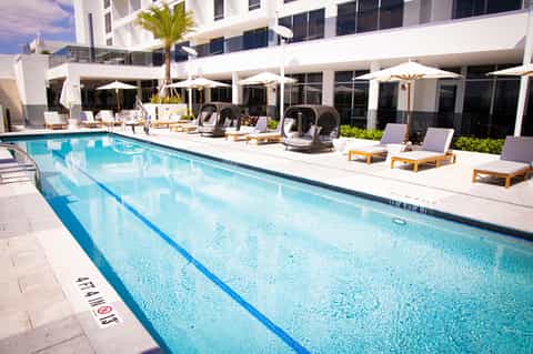 Modern resort lap pool with loungers, umbrellas, palm tree, and white building facade