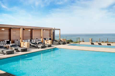 Oceanfront pool with turquoise water, modern lounge chairs, and wooden cabanas with ocean view backdrop