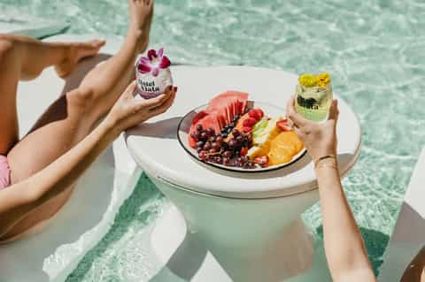 Guest holding drinks and tropical fruit plate while relaxing by turquoise water at a resort