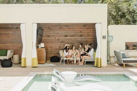 Modern pool cabana with white curtains, wooden interior, and guests relaxing by the water feature