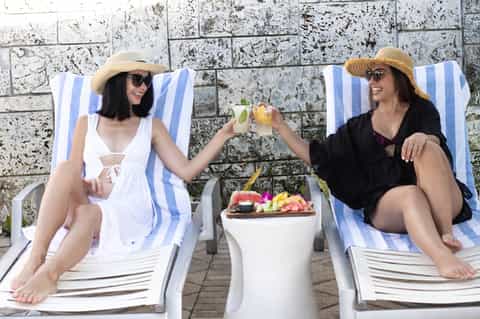 Two women toasting drinks in lounge chairs against weathered stone wall background