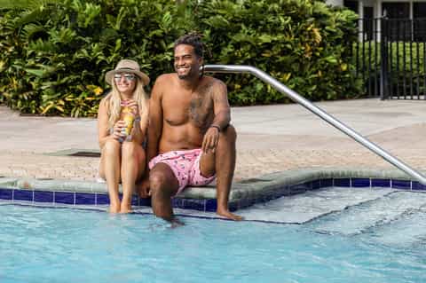 Couple sitting poolside on spa stairs holding tropical drinks near landscaped resort