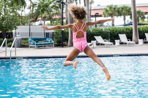 Young child in pink swimsuit jumping into resort swimming pool near palm trees and lounge seating