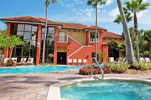 Resort exterior with red building, main pool, scattered lounge chairs, and palm trees under blue sky