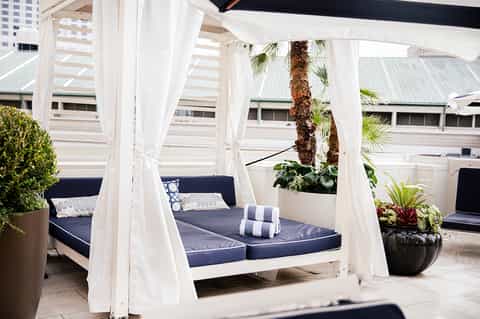 Outdoor cabana bed with white sheer curtains, navy cushions, and tropical plants on a terrace